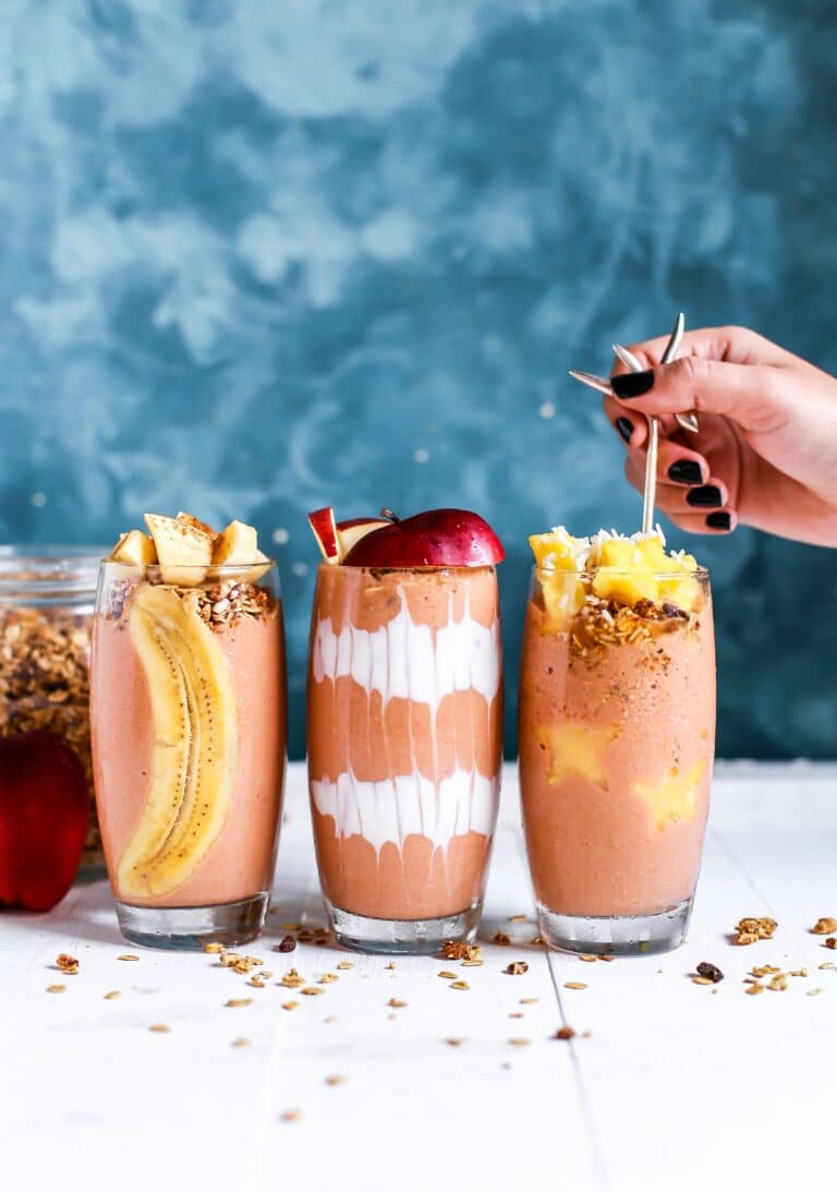Three Queen Garnet spiced apple & plum smoothie's in glasses sitting on a wooden table, with a hand putting a straw into a glass